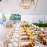 Elegant reception tent with long farm table, white chairs, amber glassware, and wicker pendant lights