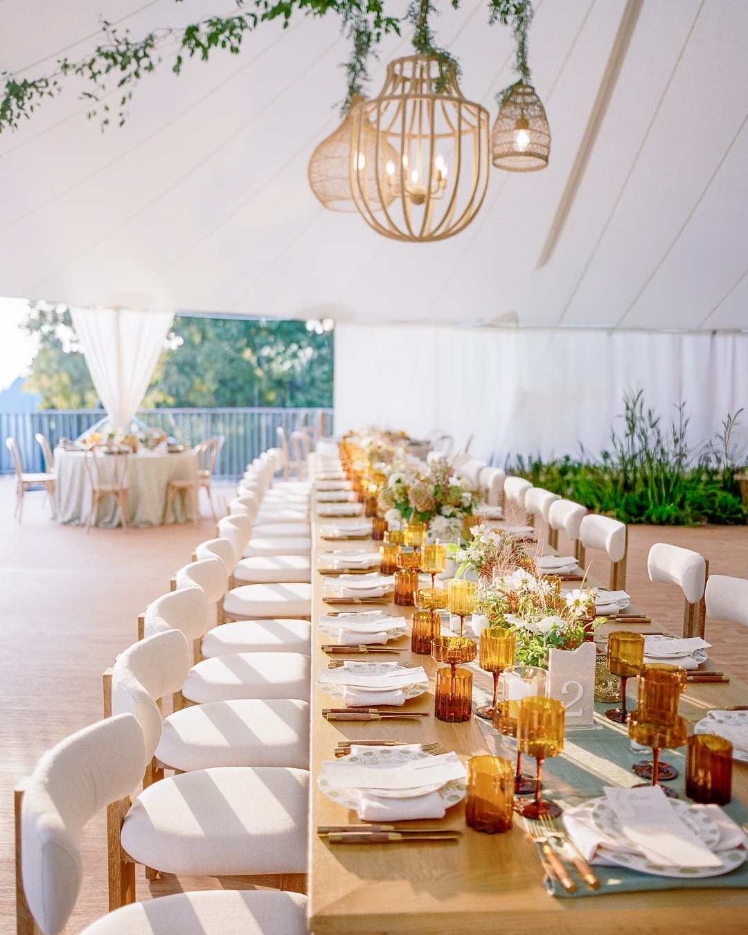 Elegant reception tent with long farm table, white chairs, amber glassware, and wicker pendant lights