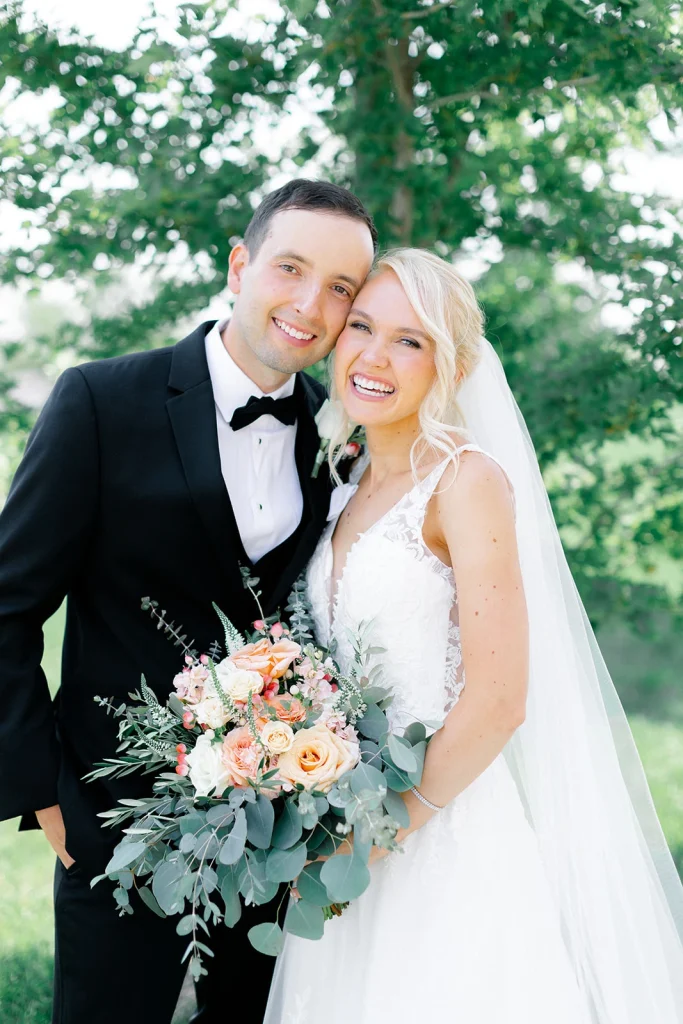 Smiling bride and groom holding peach and eucalyptus bouquet in outdoor garden setting