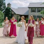 Bride in white gown with bridesmaids in pink and magenta dresses walking on gravel driveway at estate venue