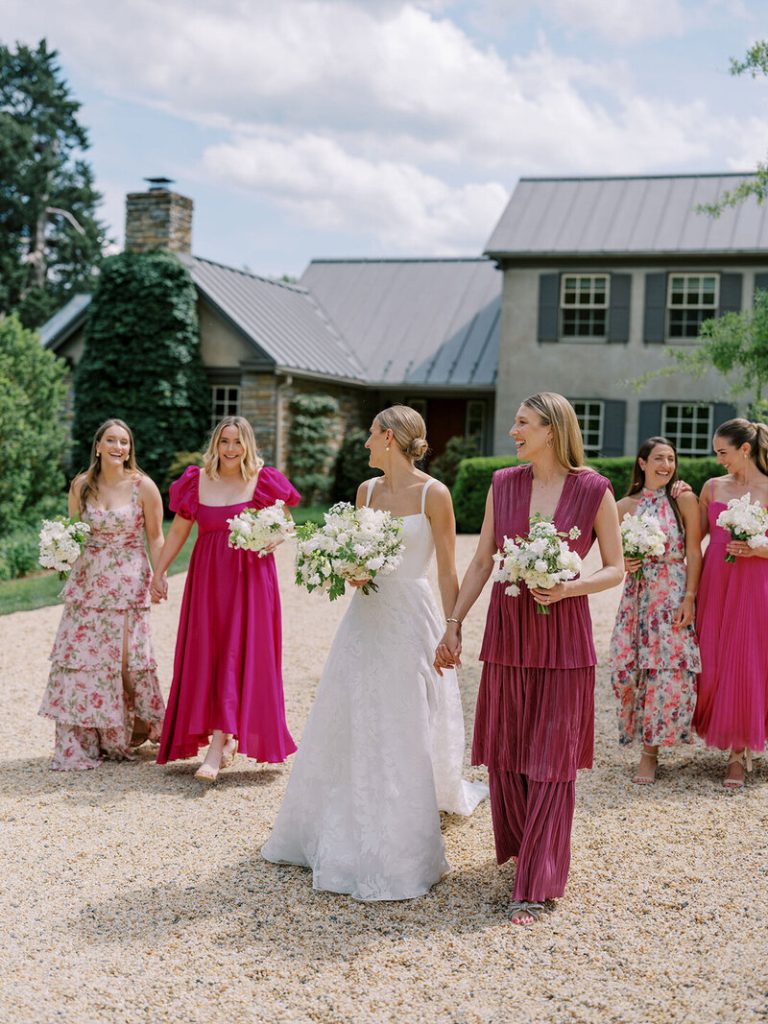Bride in white gown with bridesmaids in pink and magenta dresses walking on gravel driveway at estate venue