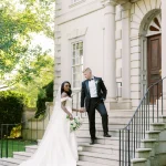 Bride and groom descending grand stone staircase of elegant white colonial building with columns