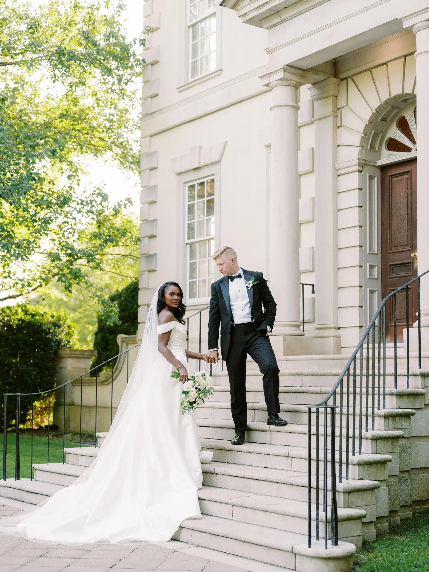 Bride and groom descending grand stone staircase of elegant white colonial building with columns