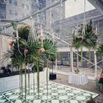 Urban rooftop wedding venue with clear tent, tropical palm arrangements, macramé chandeliers, and green geometric patterned dance floor