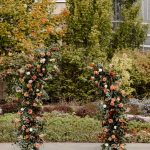 Tall floral columns with coral roses, eucalyptus, and autumn foliage framing outdoor ceremony space