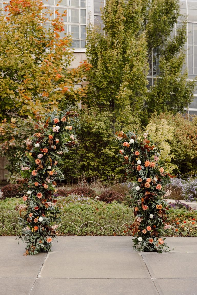 Tall floral columns with coral roses, eucalyptus, and autumn foliage framing outdoor ceremony space