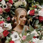 Bride with elegant updo and makeup surrounded by lush floral arrangement of roses and anemones