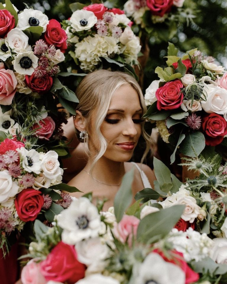 Bride with elegant updo and makeup surrounded by lush floral arrangement of roses and anemones