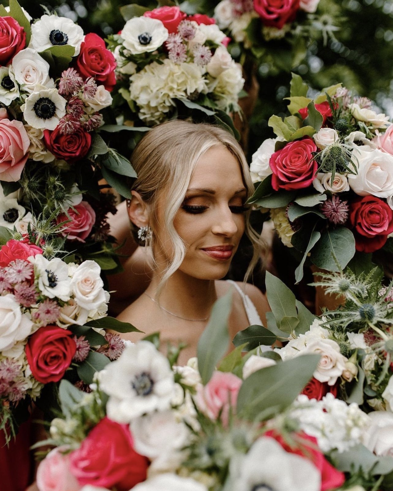 Bride with elegant updo and makeup surrounded by lush floral arrangement of roses and anemones