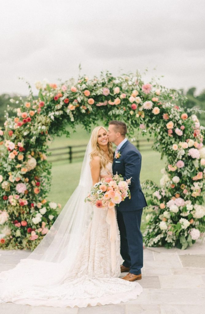 Bride and groom posing beneath a circular floral arch adorned with pink, peach, and white flowers at an outdoor Northern Virginia wedding