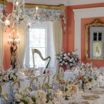 Grand banquet table with abundant pastel floral arrangements, crystal chandelier, and numbered table card in palatial coral-walled room