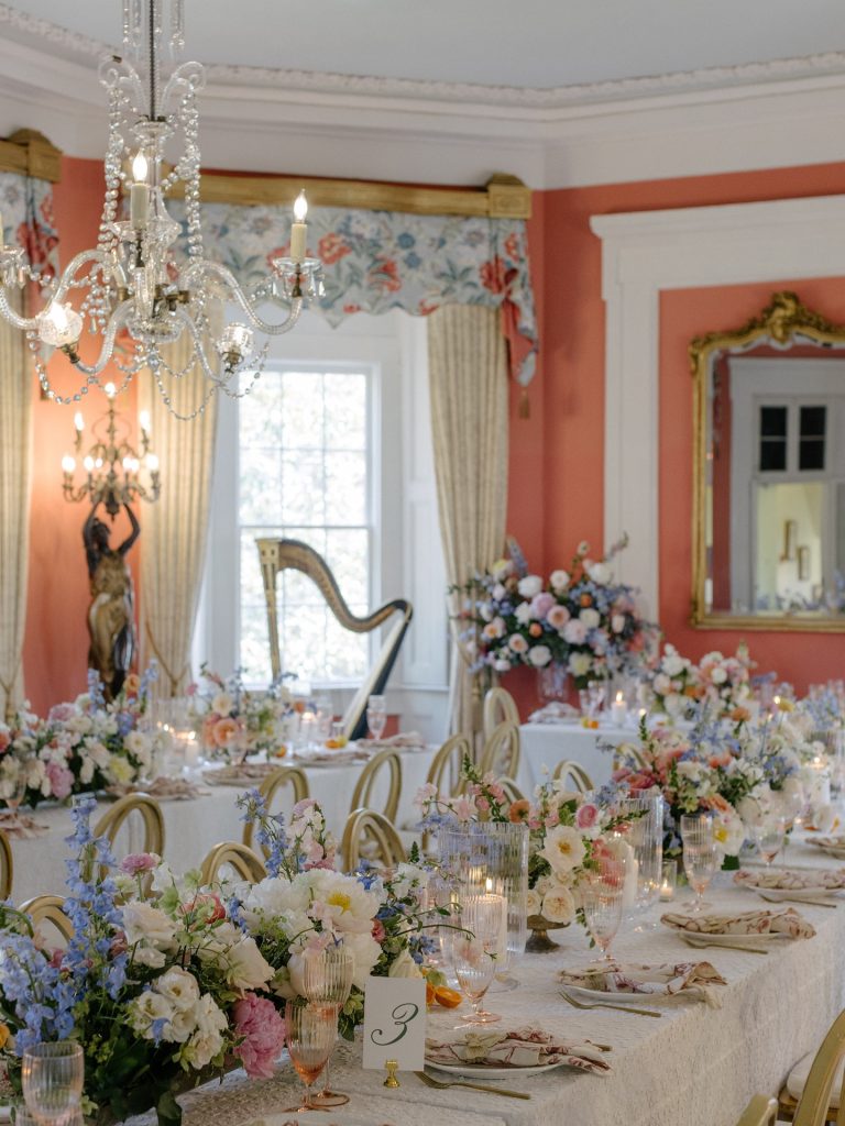 Grand banquet table with abundant pastel floral arrangements, crystal chandelier, and numbered table card in palatial coral-walled room