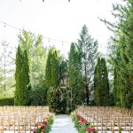 Outdoor wedding ceremony with white aisle, gold chiavari chairs, geometric floral arch with pink and coral roses, and tall evergreen trees with string lights overhead