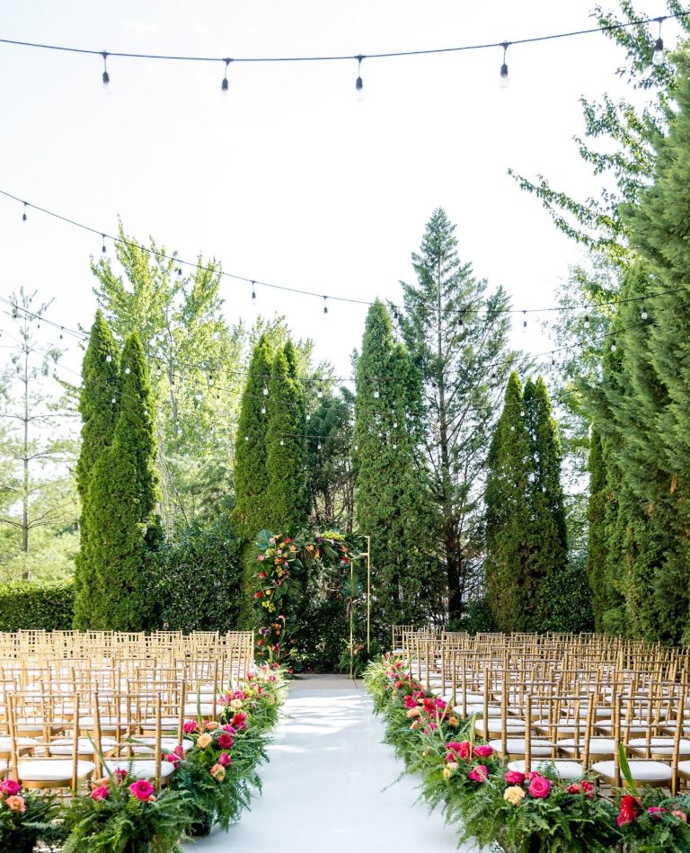Outdoor wedding ceremony with white aisle, gold chiavari chairs, geometric floral arch with pink and coral roses, and tall evergreen trees with string lights overhead