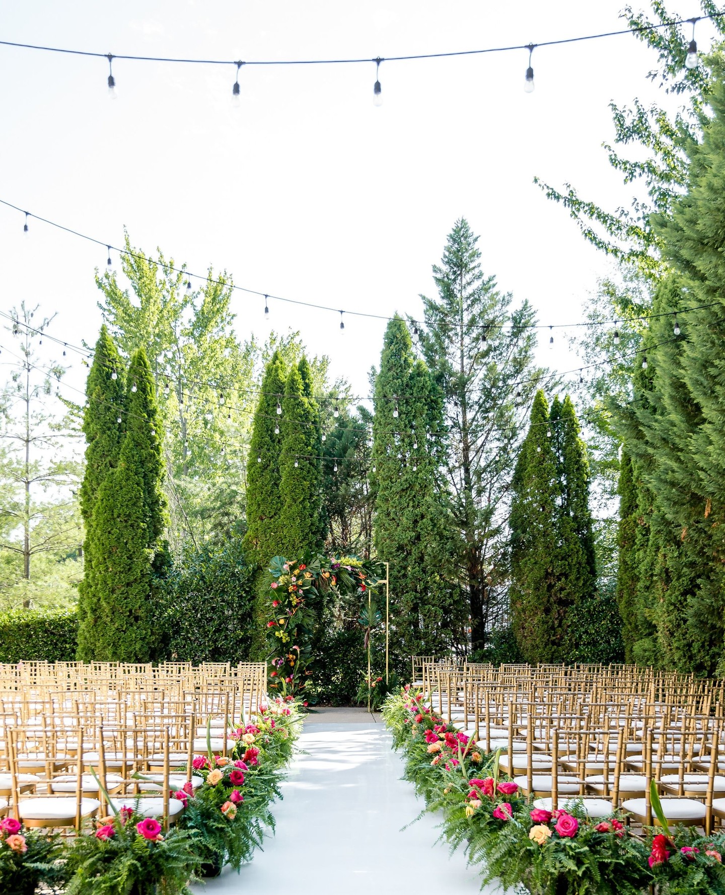 Outdoor wedding ceremony with white aisle, gold chiavari chairs, geometric floral arch with pink and coral roses, and tall evergreen trees with string lights overhead