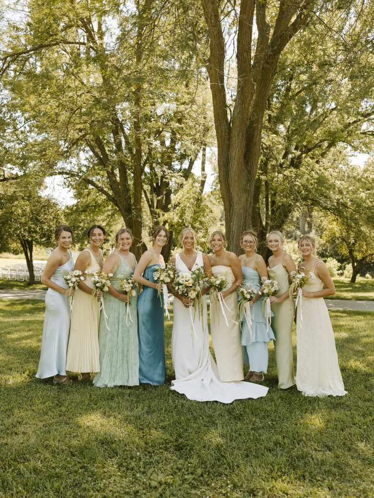 Bride and bridesmaids in mismatched pastel gowns holding white and cream bouquets under trees in Nebraska park