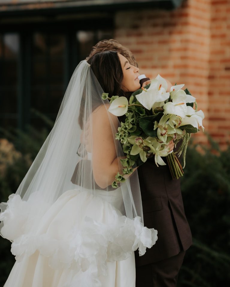 Bride in white ball gown and veil smelling white calla lily bouquet outdoors at brick building