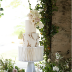 Three-tier white wedding cake with gold accents, ruffled bottom tier, and blush sugar flowers displayed in garden setting