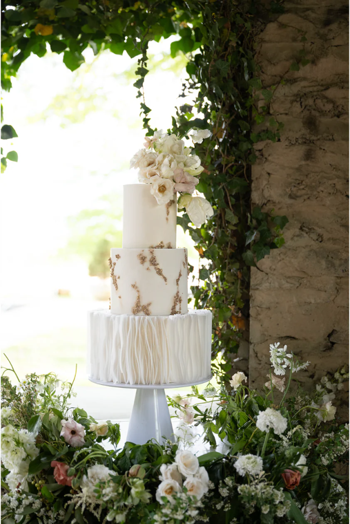Three-tier white wedding cake with gold accents, ruffled bottom tier, and blush sugar flowers displayed in garden setting