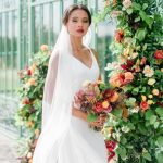 Bride in white gown holding colorful bouquet beside floral arch at greenhouse wedding venue