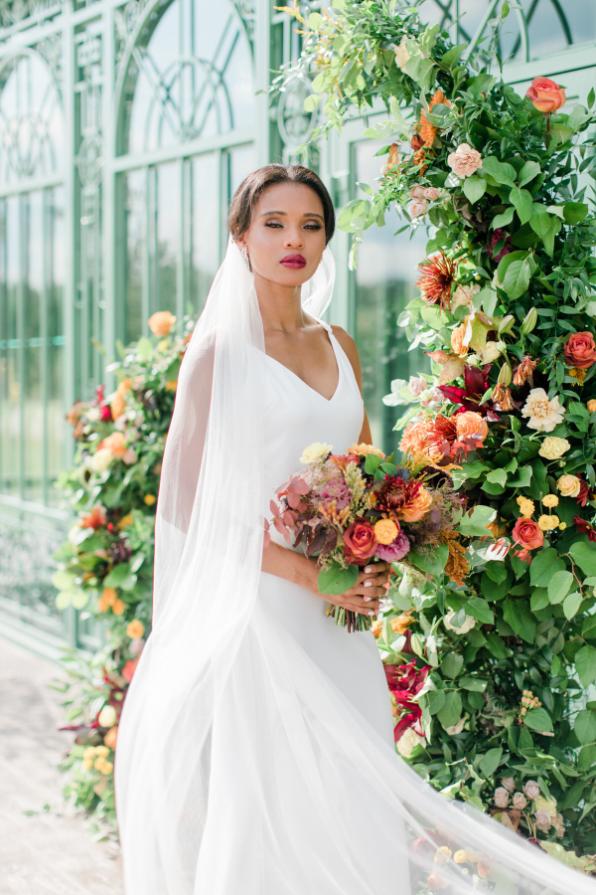 Bride in white gown holding colorful bouquet beside floral arch at greenhouse wedding venue