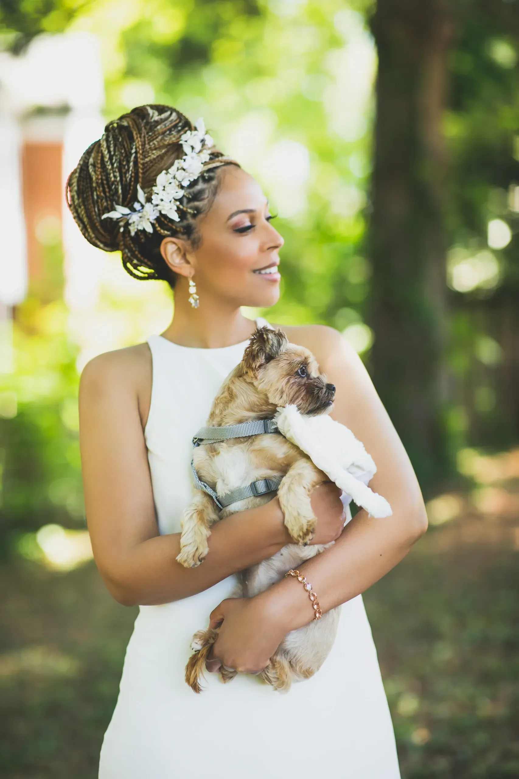 Bride with braided updo and floral crown holding small dog outdoors in natural light