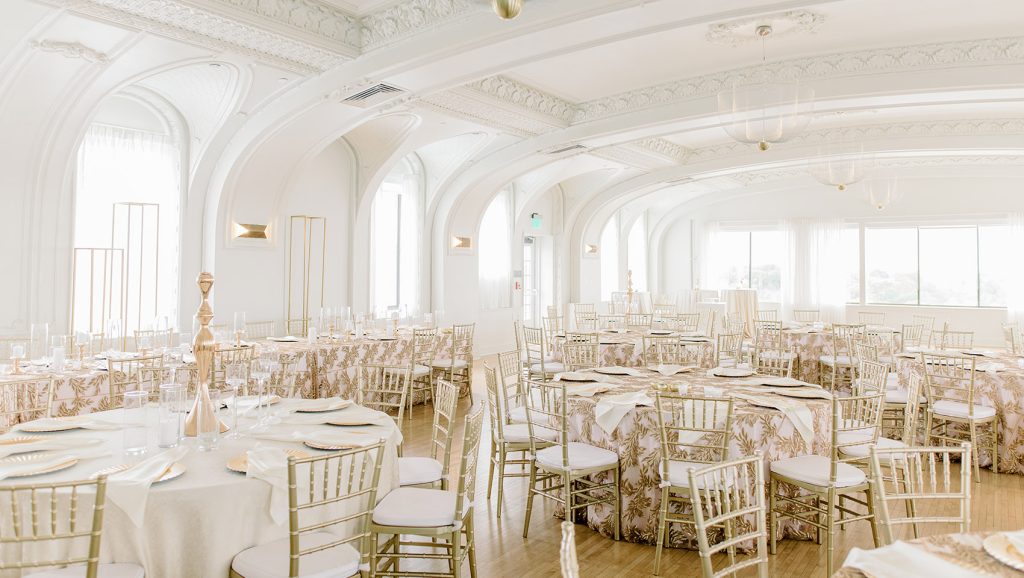 Spacious wedding reception ballroom with white arched columns, gold chiavari chairs, and embroidered table linens