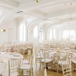 Spacious wedding reception ballroom with white arched columns, gold chiavari chairs, and embroidered table linens