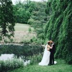 Bride and groom embracing by a tranquil pond under cascading willow trees in a lush garden setting