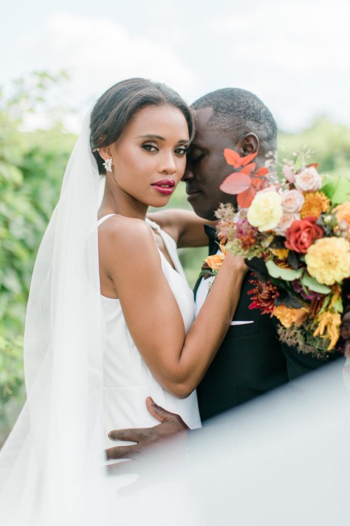 Bride with elegant updo and burgundy lipstick poses with groom holding vibrant fall bouquet