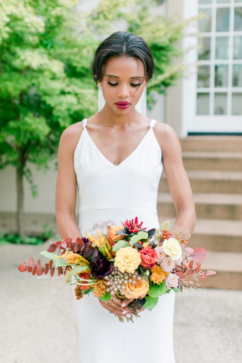 Bride in minimalist white V-neck gown holding colorful fall bouquet with peach roses, eucalyptus, and burgundy blooms