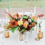 Autumn wedding centerpiece with orange and burgundy flowers, white candles, and amber glassware