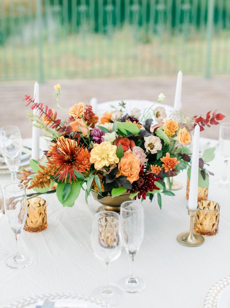 Autumn wedding centerpiece with orange and burgundy flowers, white candles, and amber glassware