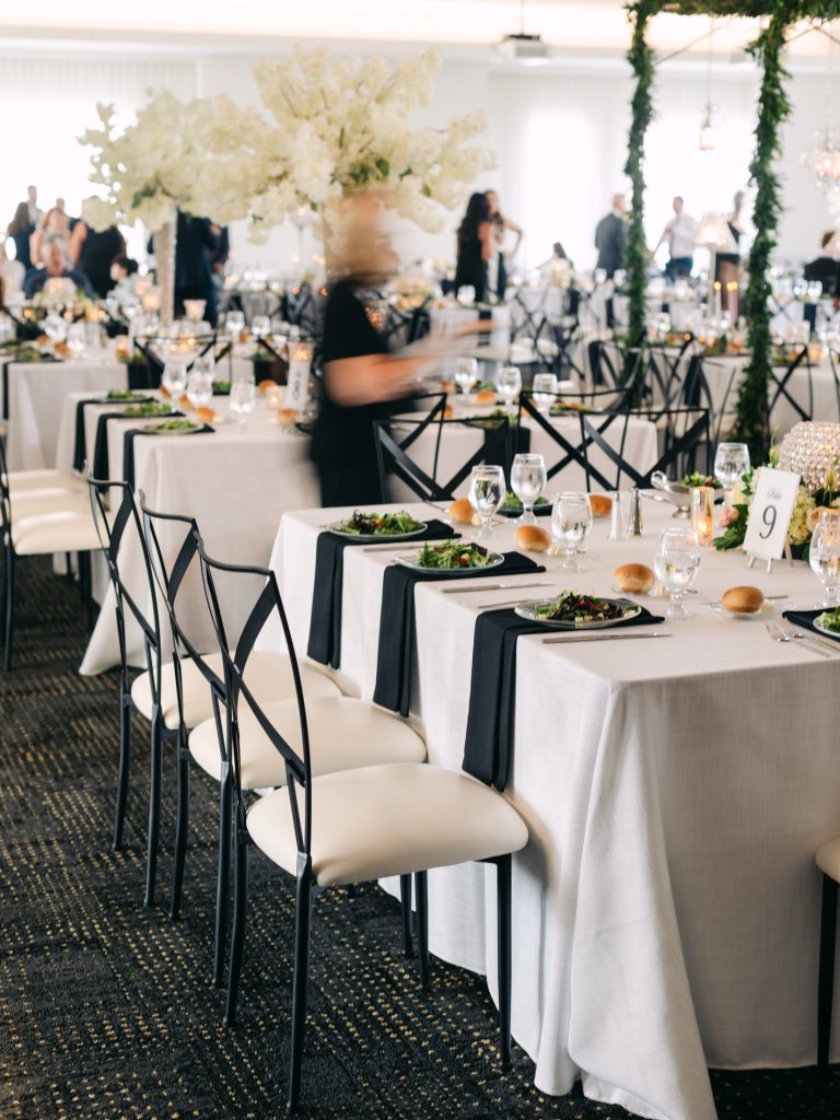 Elegant wedding reception with white linens, black napkins, tall floral centerpieces, and modern white chairs