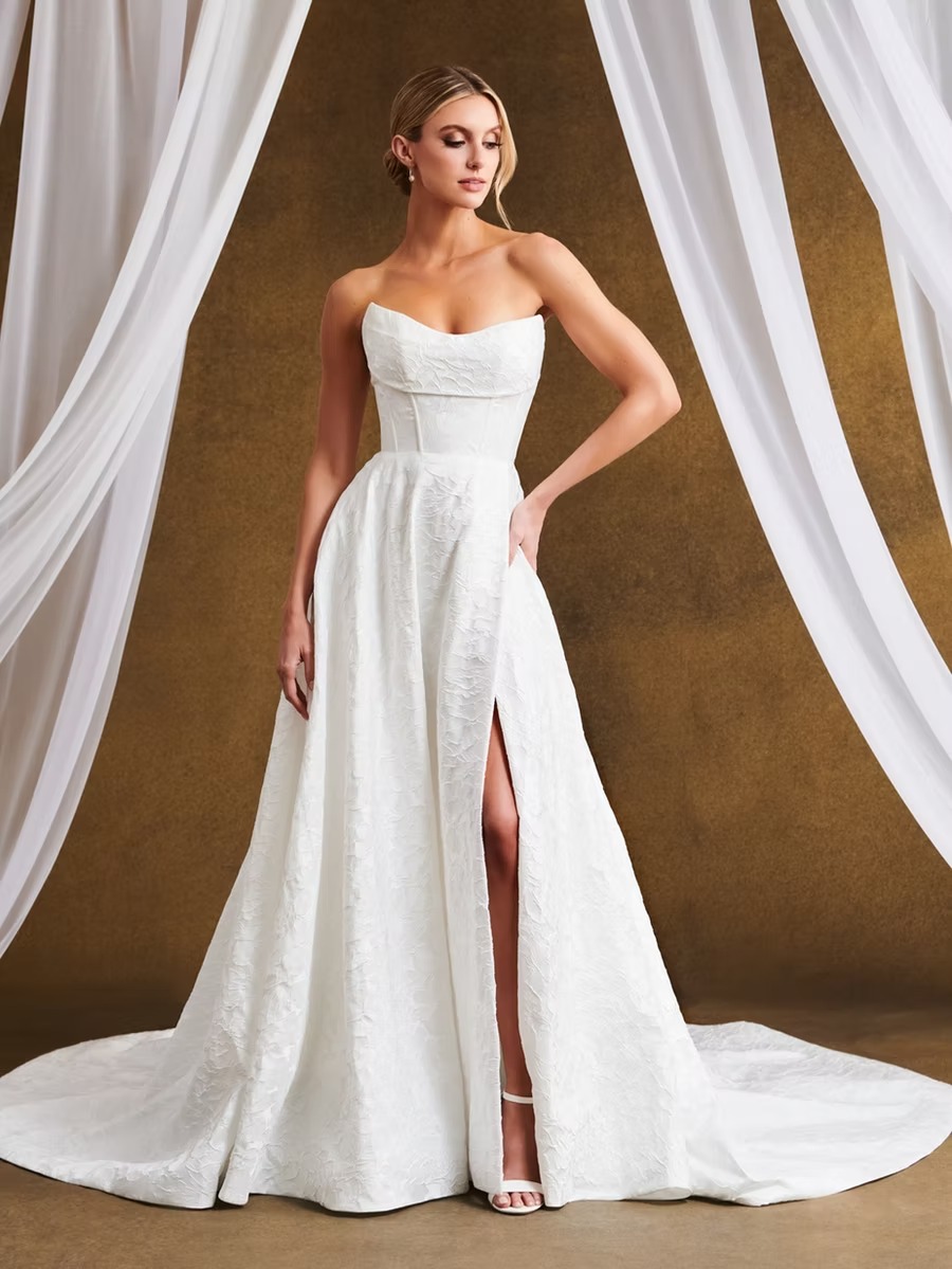 Bride in strapless A-line wedding gown with high front slit and cathedral train, framed by white draping against gold backdrop