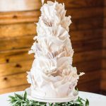 White tiered wedding cake with textured fondant ruffles surrounded by green foliage wreath on rustic wood background