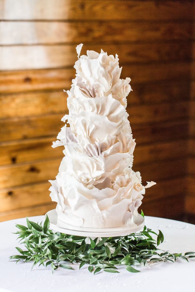 White tiered wedding cake with textured fondant ruffles surrounded by green foliage wreath on rustic wood background