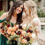 Bride and bridesmaid laughing together holding matching bouquets of peach roses, burgundy dahlias, and greenery