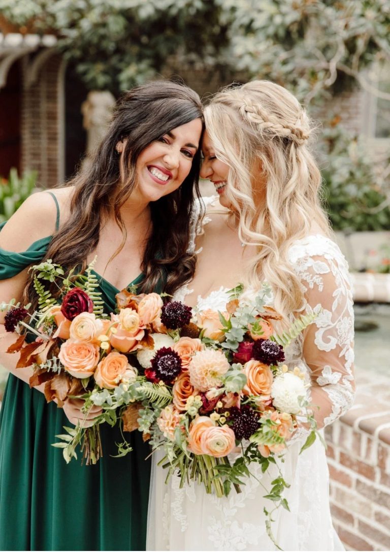 Bride and bridesmaid laughing together holding matching bouquets of peach roses, burgundy dahlias, and greenery