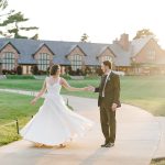 Bride twirls in flowing white dress with groom on path outside clubhouse at sunset