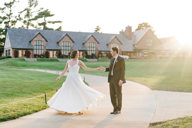Bride twirls in flowing white dress with groom on path outside clubhouse at sunset