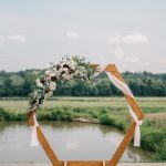 Wooden hexagon ceremony arch decorated with white and burgundy florals, eucalyptus, and blush fabric beside pond