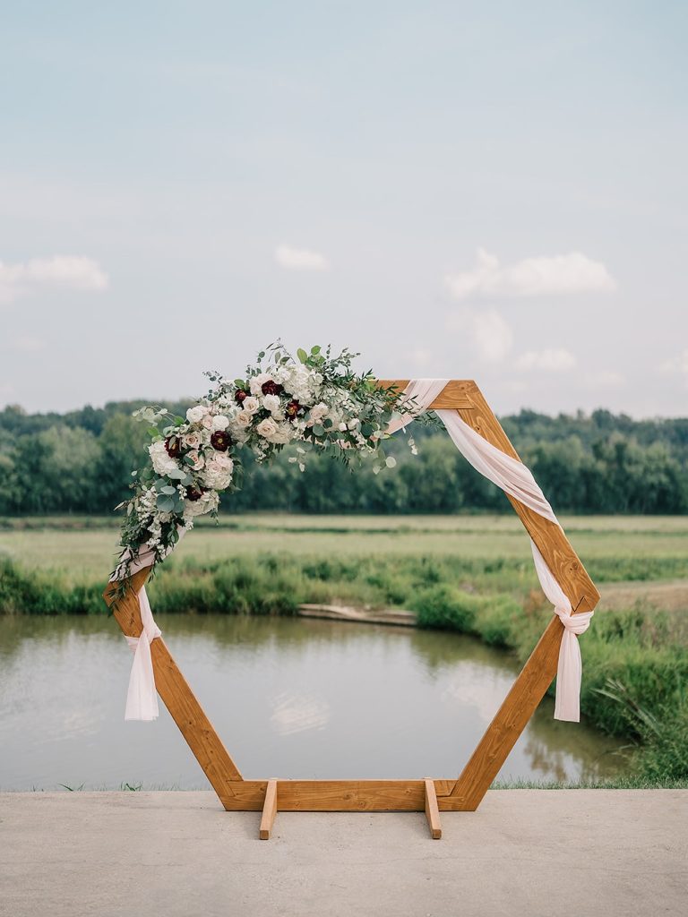 Wooden hexagon ceremony arch decorated with white and burgundy florals, eucalyptus, and blush fabric beside pond