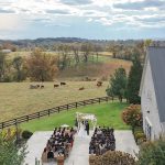 Aerial view of outdoor wedding ceremony at farm venue with guests seated, white barn, and cattle in rolling pastures beyond