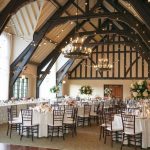 Wedding reception setup in barn venue featuring dramatic exposed beams, bistro lighting, and romantic floral arrangements