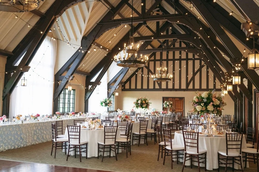 Wedding reception setup in barn venue featuring dramatic exposed beams, bistro lighting, and romantic floral arrangements