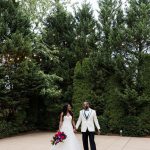Bride in white ball gown and groom in white jacket holding hands on stone patio with vibrant red and purple bouquet under string lights and trees