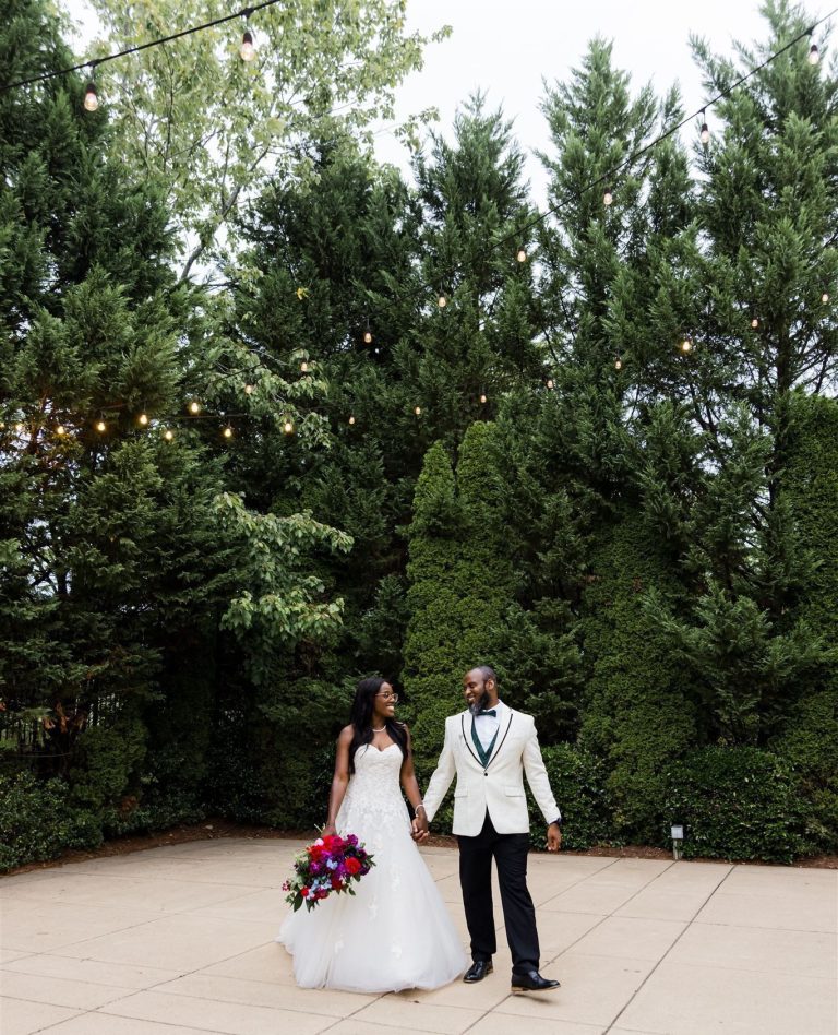 Bride in white ball gown and groom in white jacket holding hands on stone patio with vibrant red and purple bouquet under string lights and trees