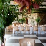 Garden-inspired reception with stone wall backdrop, pampas grass installation, cane-back chairs, and checkerboard floor