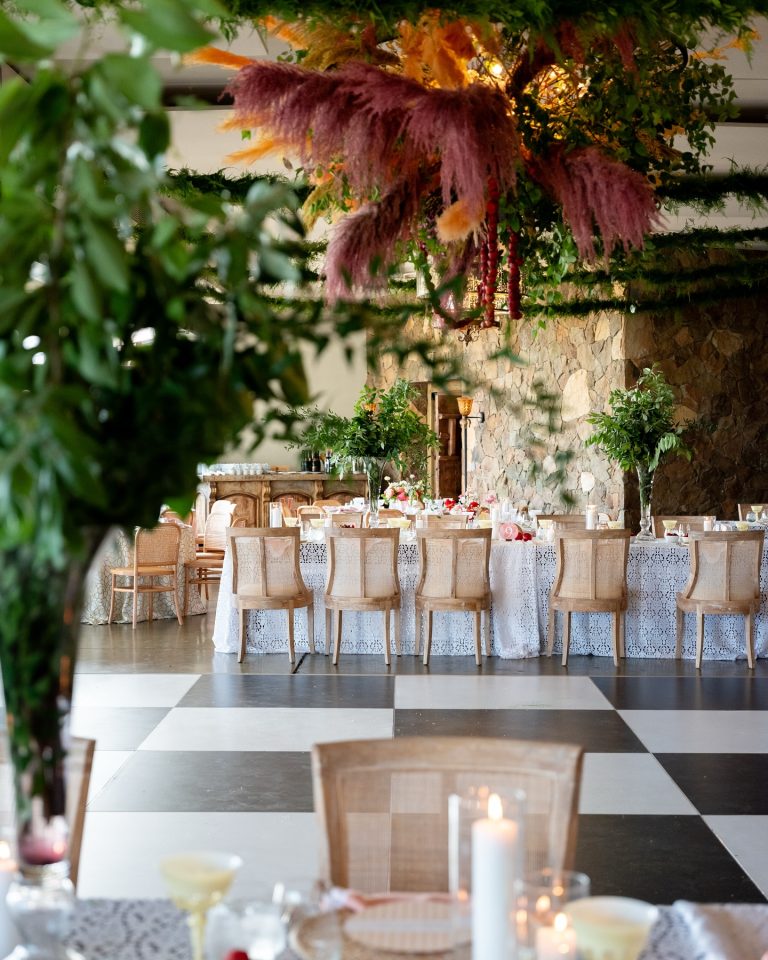 Garden-inspired reception with stone wall backdrop, pampas grass installation, cane-back chairs, and checkerboard floor